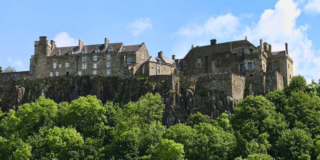 Stirling Castle perched dramatically atop a sheer volcanic rock cliff face surrounded by lush green treetops under a bright blue sky